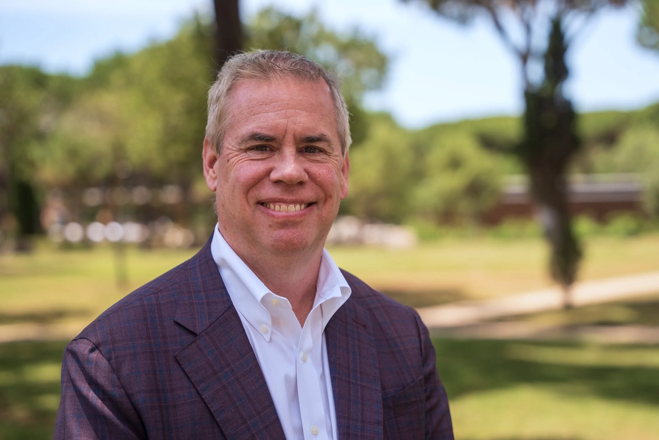 Portrait of a middle-aged man with short blonde hair, wearing a dark plaid blazer and white shirt, smiling outdoors in a park with trees and a blue sky in the background.