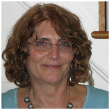 A smiling woman with curly brown hair, glasses, and a turquoise top, standing indoors near a staircase.