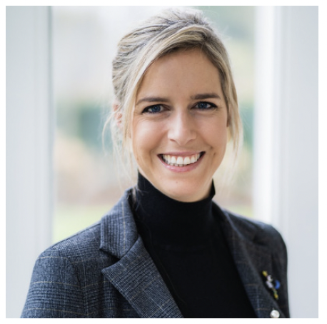 Portrait of a woman with blonde hair in a ponytail, wearing a black turtleneck and a gray blazer, smiling in front of a window.