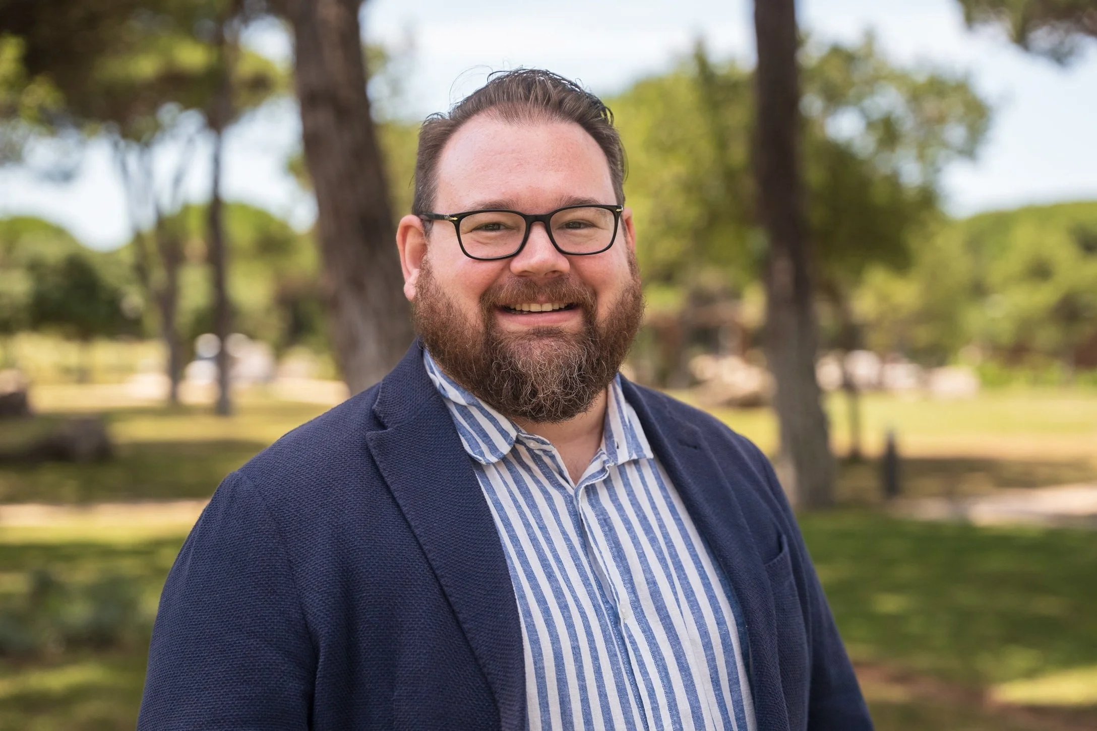 A man with glasses, a beard, and a smile, wearing a blue blazer and a striped shirt, standing outdoors in a park with trees in the background.