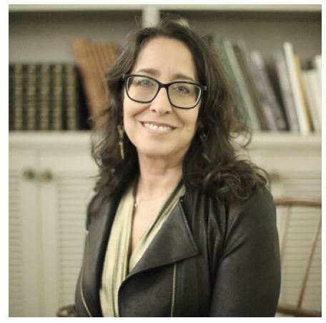 A woman with dark, wavy hair and glasses smiling, wearing a black leather jacket and a scarf, in front of a bookshelf.