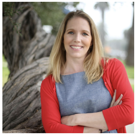 A woman with blonde hair, wearing a red cardigan and blue shirt, smiling with arms crossed outdoors near a tree.