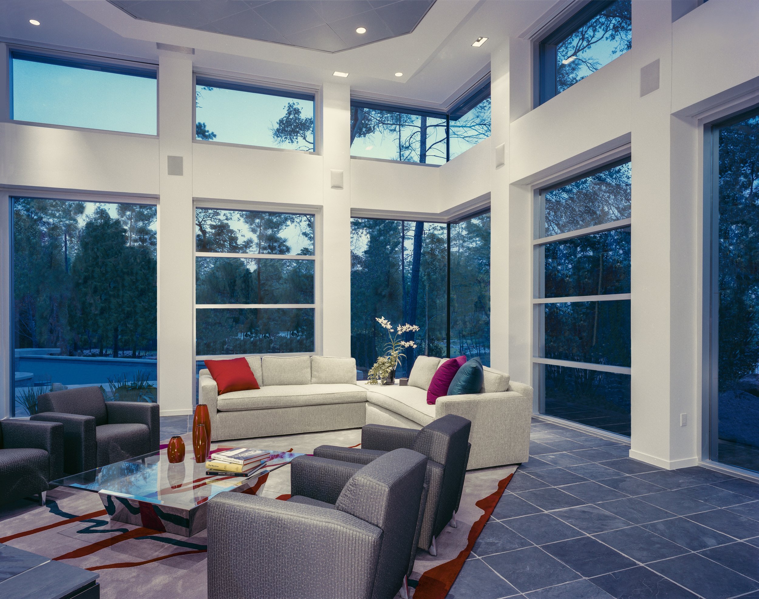 Modern living room with large floor-to-ceiling windows, white walls, custom sectional sofa with colorful pillows, grey armchairs, a glass coffee table with decorative vases, a custom area rug, and a view of trees and pool outside.