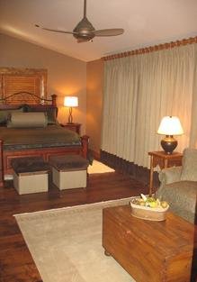 A guest bedroom with a wooden bed frame, custom curtains, and two small lamps on bedside tables, along with a cozy armchair, wooden coffee table with a tray, and a ceiling fan.