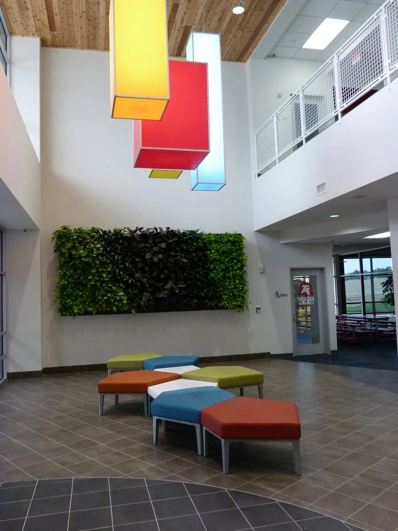 Interior of a modern building lobby with colorful hanging light fixtures, a green wall, and multi-colored seating stools arranged on tiled floors.