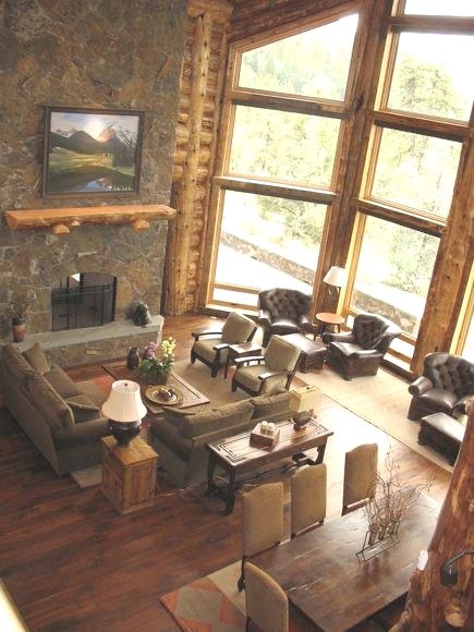 Living room in a log cabin-style house with large windows, a stone fireplace, furnishings to relax and enjoy the spectacular views, a dining table, and decorative touches including custom artwork and rugs.