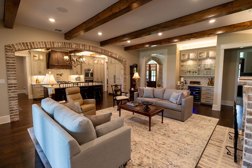 Open concept living room and kitchen with beige sofas, a stone fireplace, wooden ceiling beams, and cabinetry in the kitchen area.
