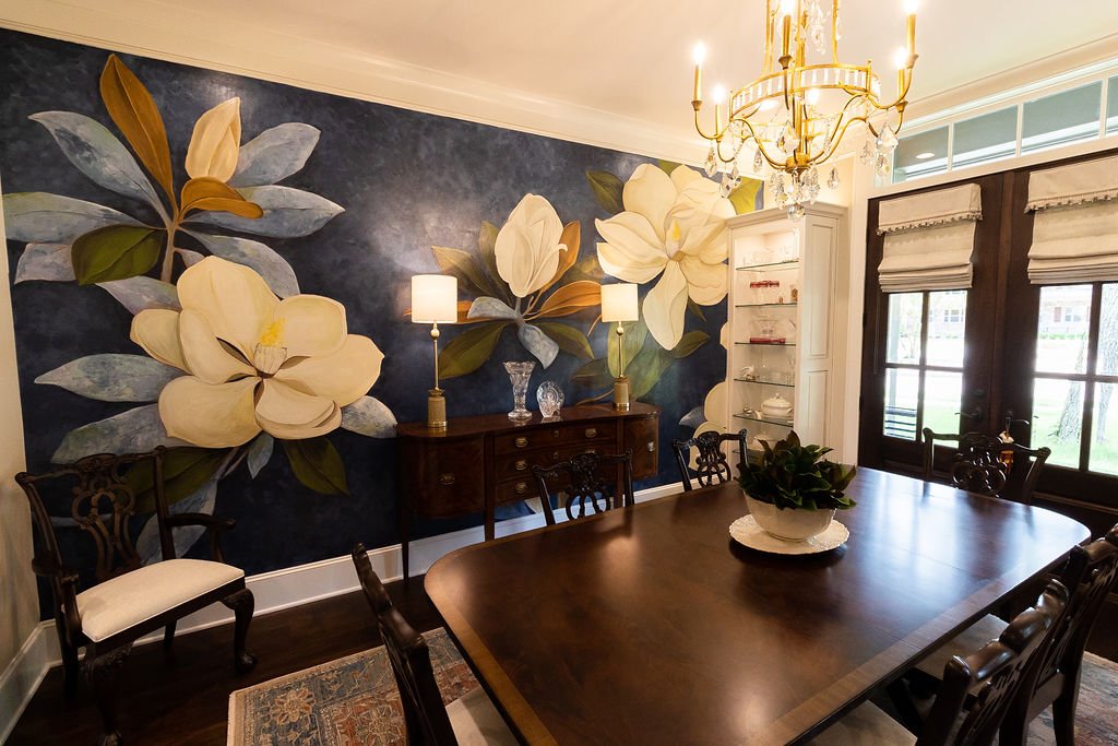 A dining room featuring a hand painted mural with white magnolia flowers and green leaves on a dark blue background. The room has a dark wood table, ornate wooden chairs, a white cabinet, a chandelier, and French doors with custom roman shades.