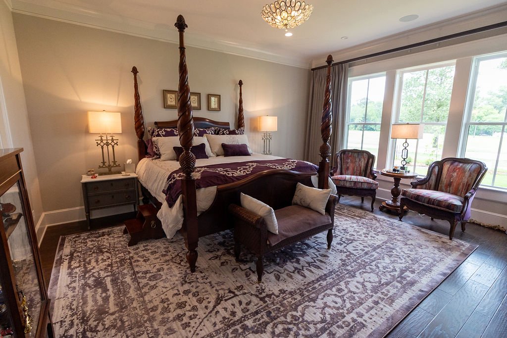 A bedroom with a four-poster bed, nightstands with lamps, two upholstered armchairs near large windows, a patterned area rug, and a small bench at the foot of the bed. Custom draperies are motorized.