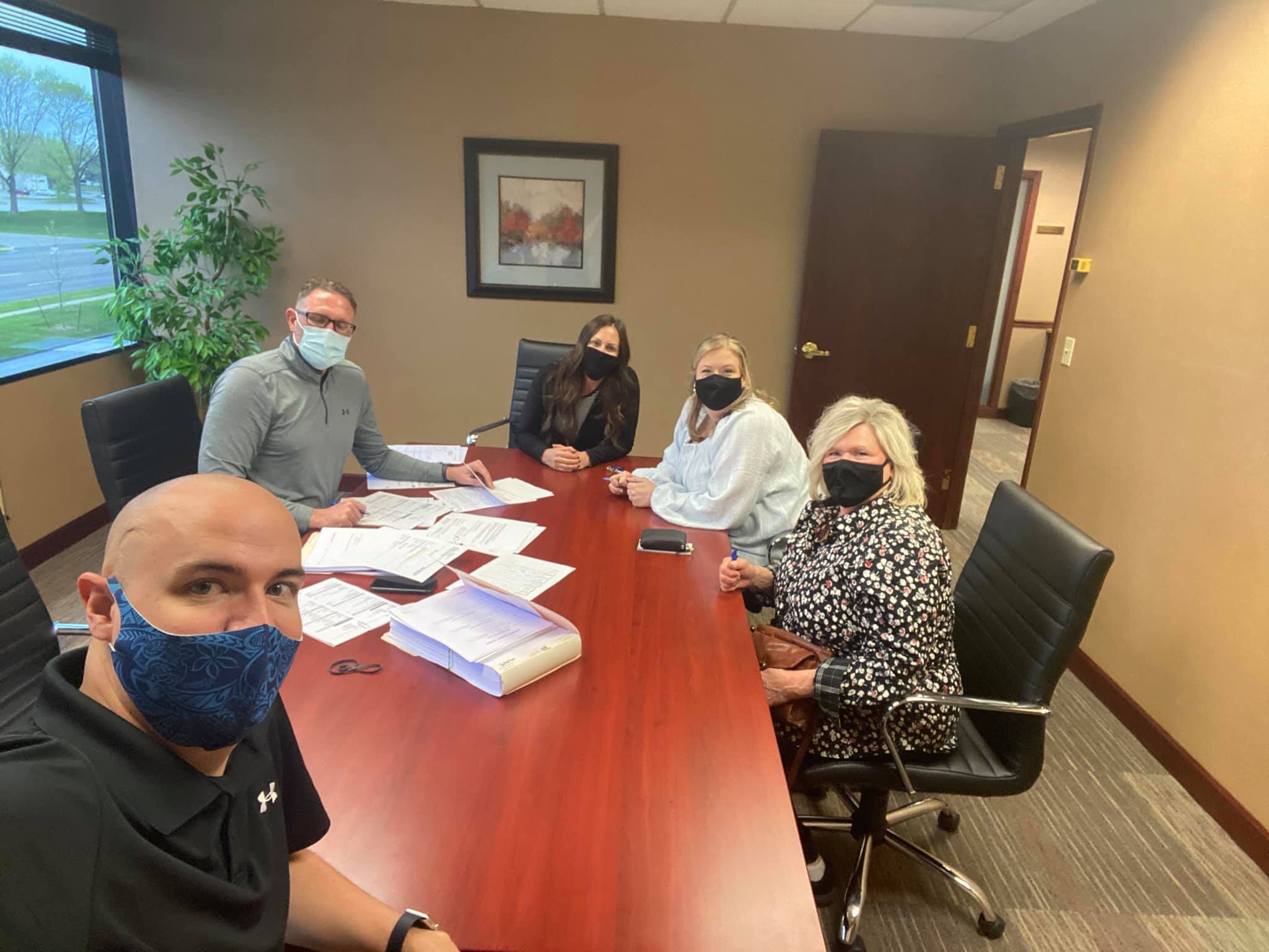 Five people, wearing face masks and smiling with their eyes, sitting around table with documents in front of them.