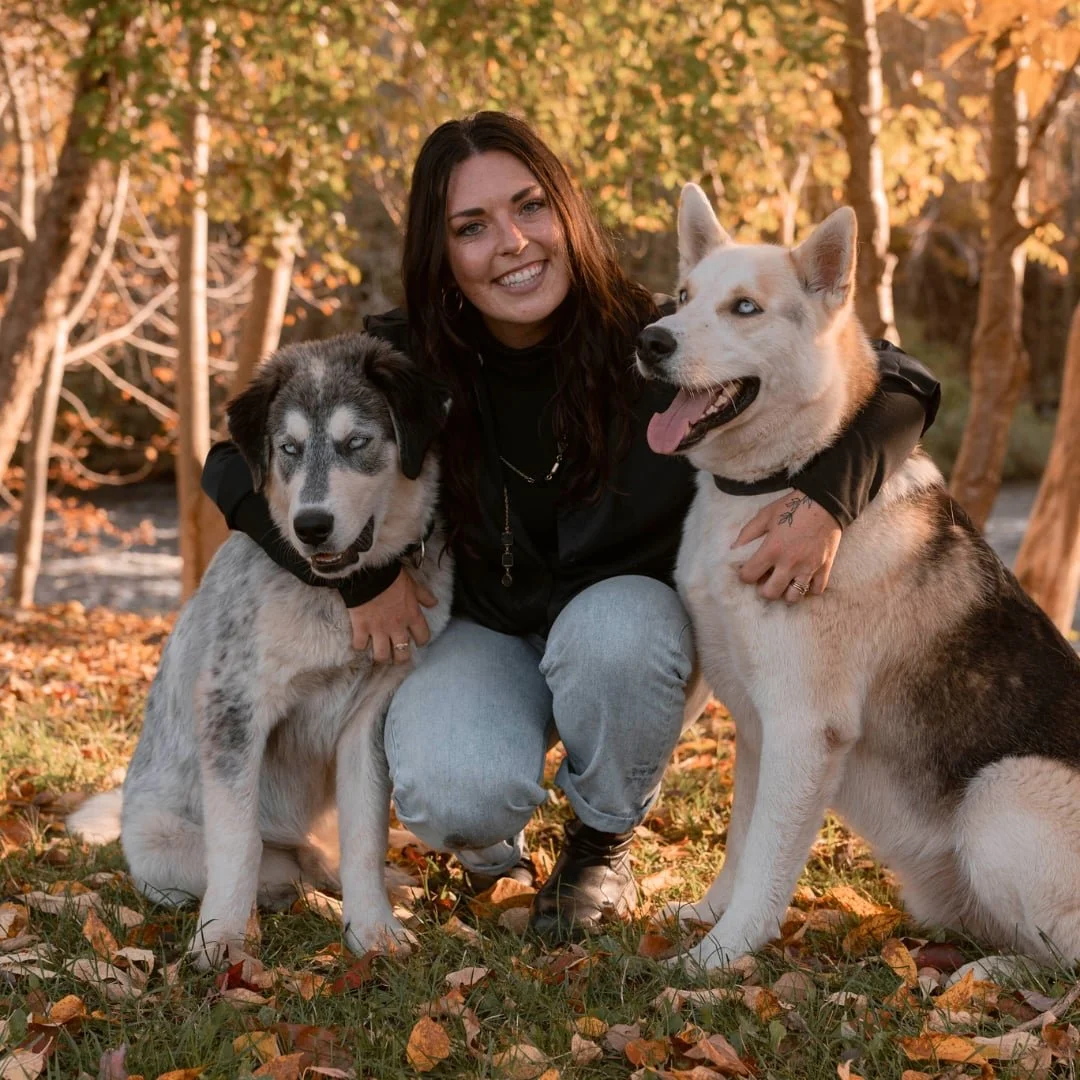 A woman smiling and crouching outdoors with two husky dogs, one black and white and the other white and brown, in an autumn setting with fall leaves and trees in the background.
