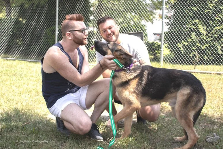 Two young white men kneel on a lawn, greeting a happy German shepherd on adoption day.
