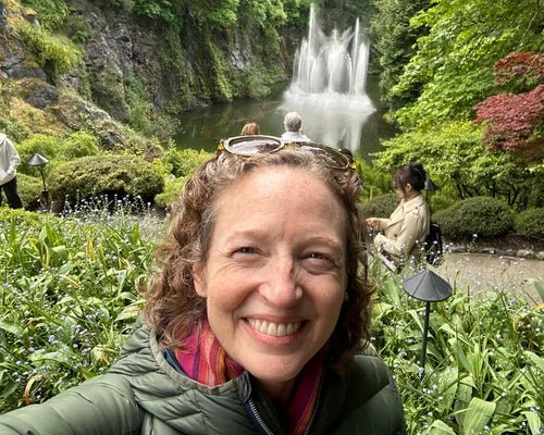 A smiling woman taking a selfie at a lush garden with a waterfall in the background.