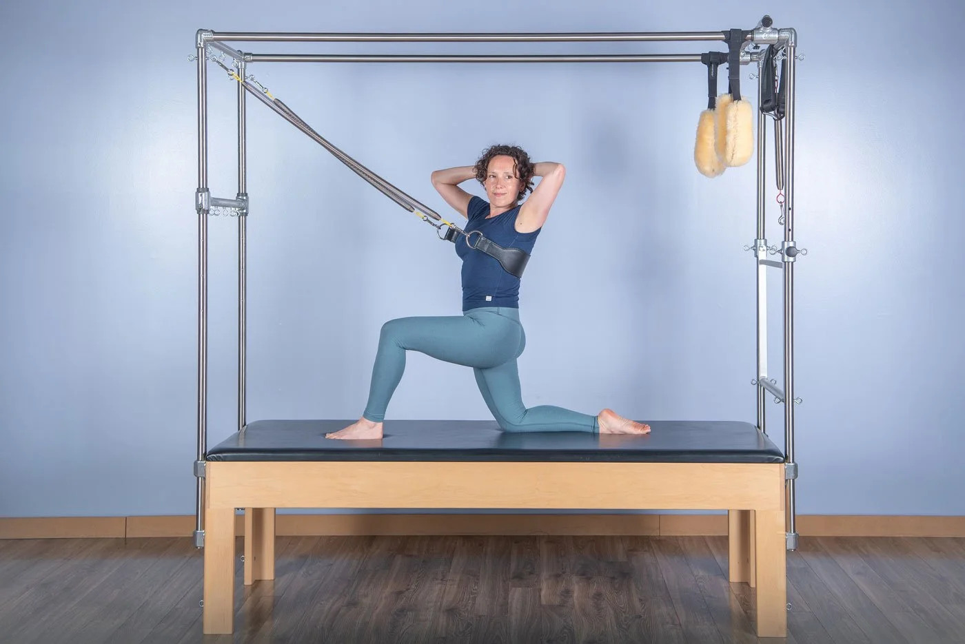 Jennifer Golden performing a Pilates exercise on a reformer machine, kneeling with one knee on the padded surface and the other foot flat, with her hands behind her head, in a fitness studio with a light blue wall.