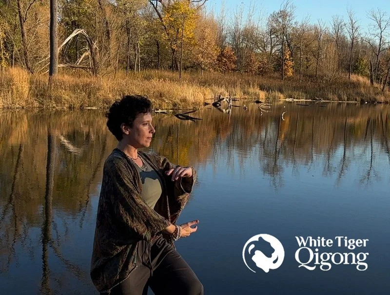 Jennifer Golden practicing Qigong by a river during autumn, with trees and leaves reflecting on the water, and the logo of White Tiger Qigong in the bottom right corner.
