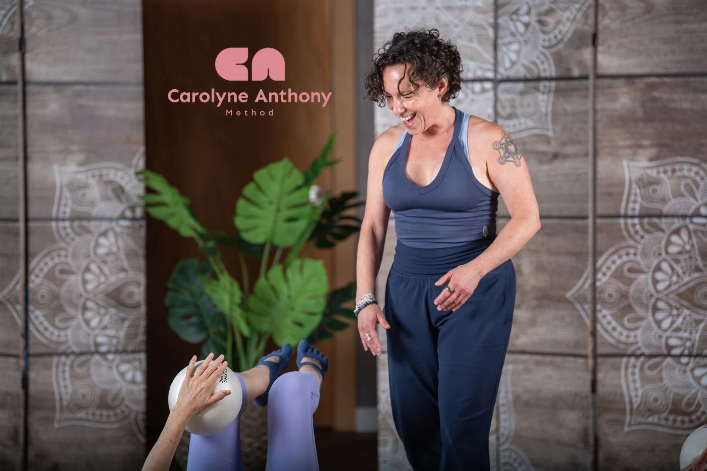 Jennifer Golden smiling and observing student’s legs using a prop during a class in studio with wooden paneling