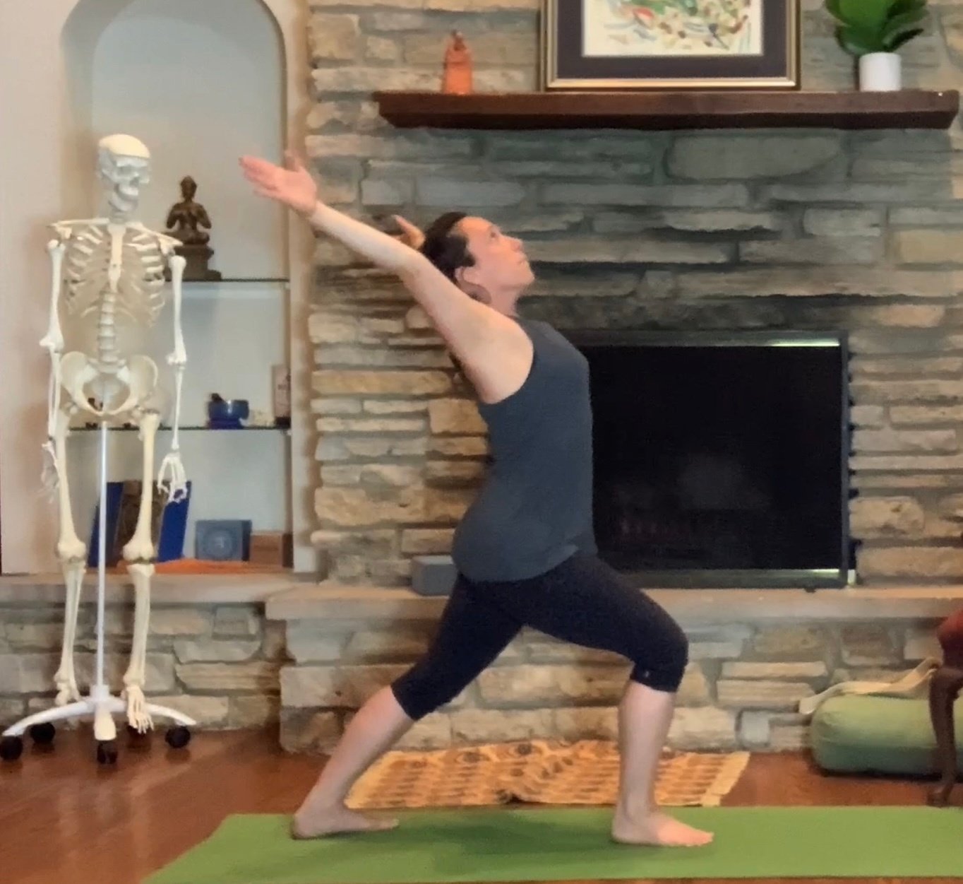 Jennifer Golden practicing pilates indoors on a green mat, in a warrior pose with arms extended upward, wearing a gray top and black leggings, in front of a stone fireplace and a decorative skeleton model.