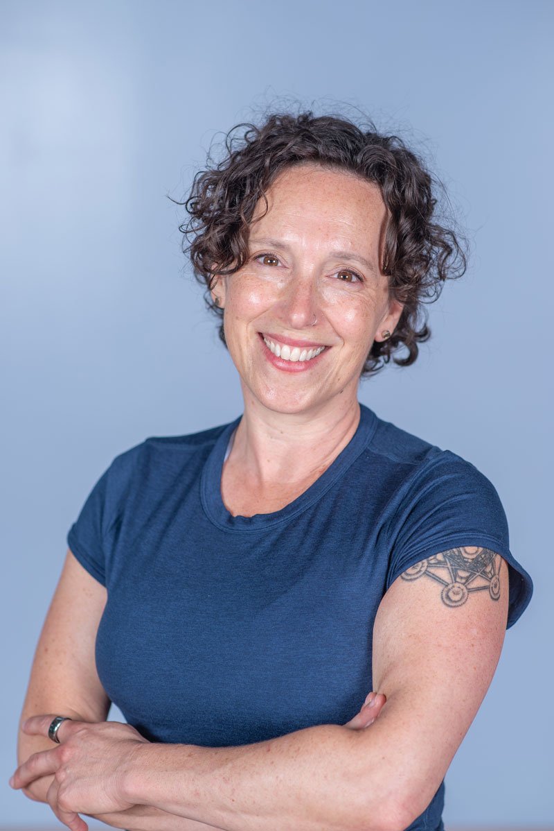 Jennifer Golden wearing a blue t-shirt, showing a tattoo on her upper arm, against a light blue background.