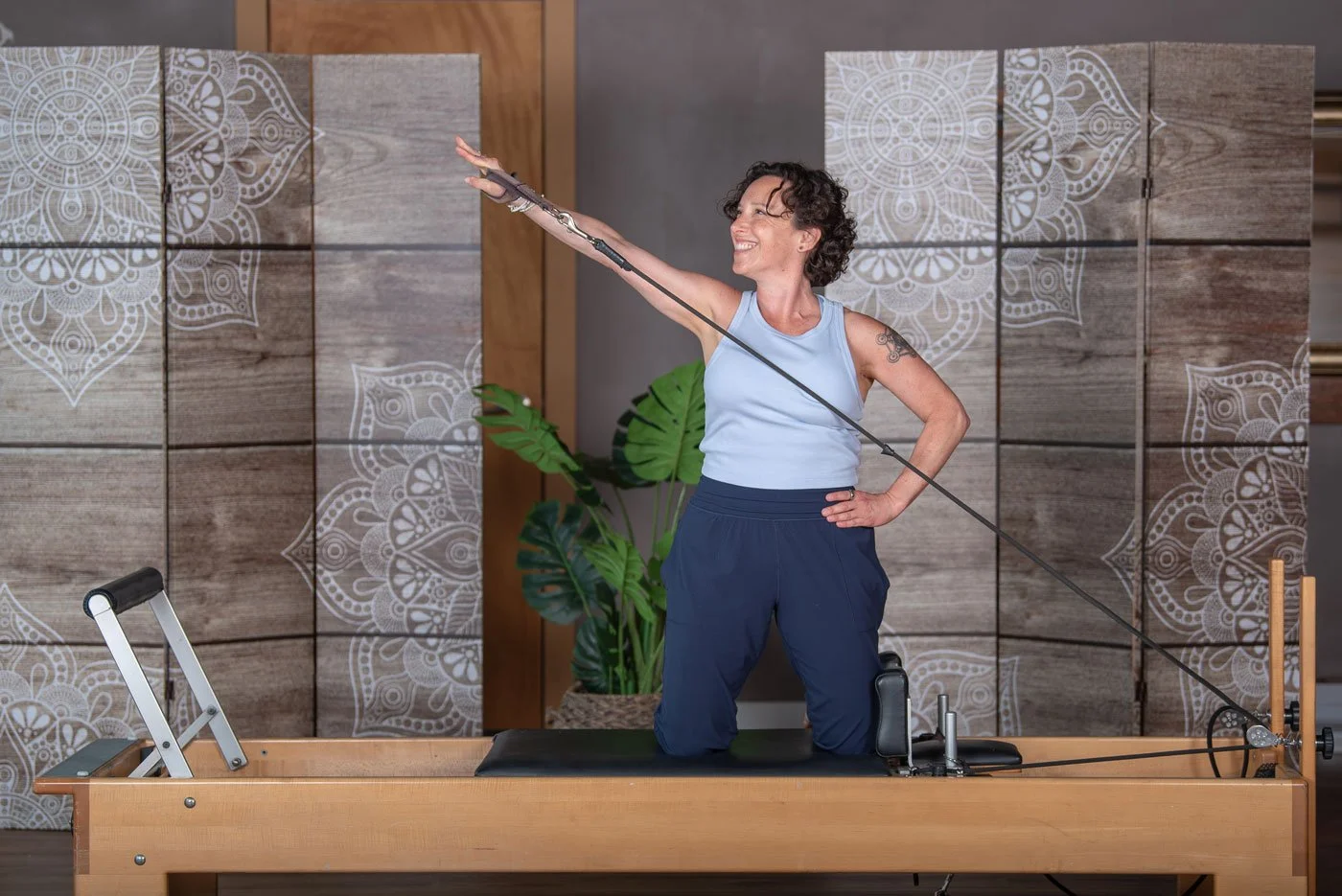 Jennifer Golden, wearing a light blue sleeveless top and dark blue pants, is kneeling on a Pilates reformer machine, stretching one arm upward and outward, in a Pilates studio with decorative wooden panels and potted plant in the background.