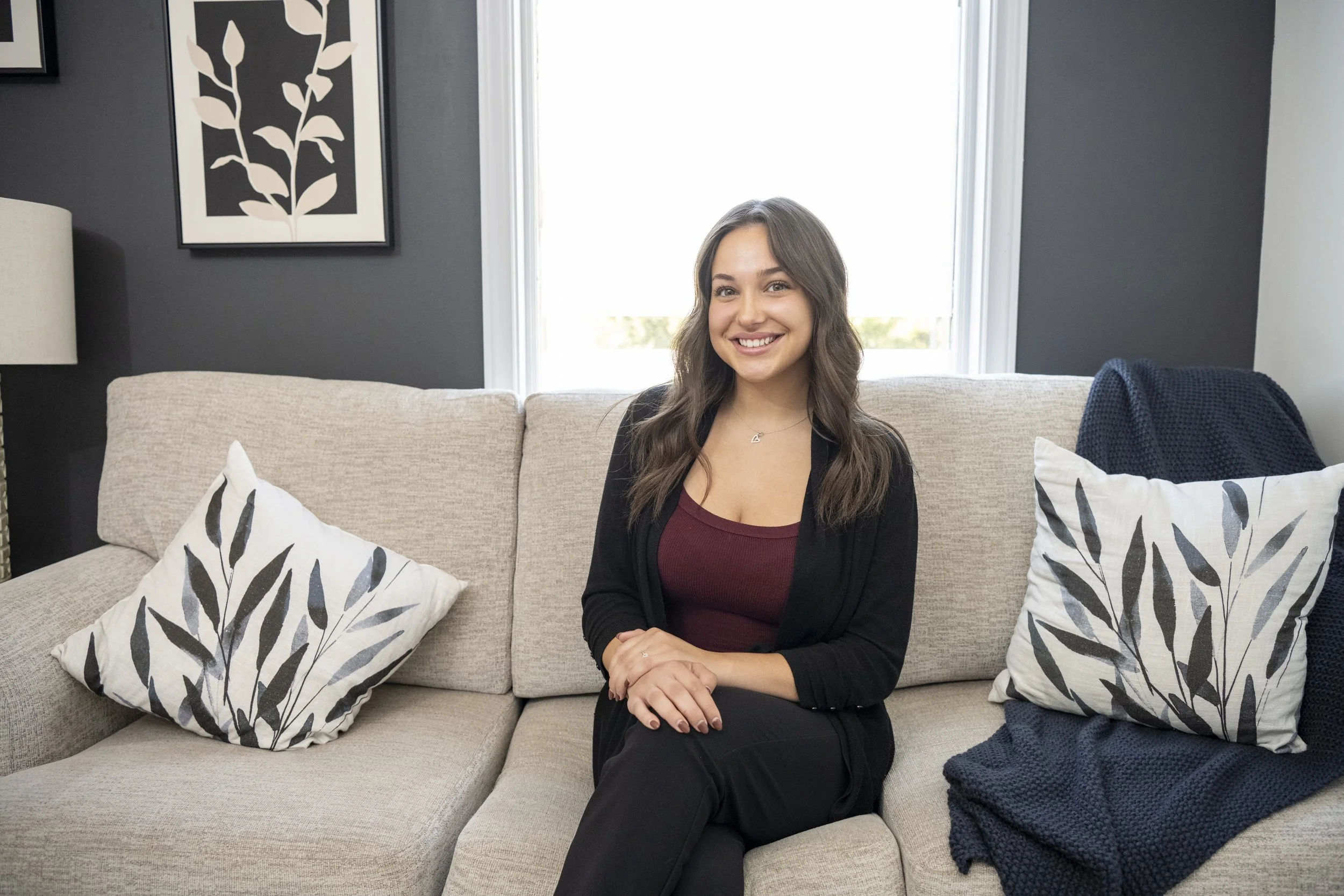 Grace McGrath smiles and sits on a grey couch with legs crossed and hands gently resting on her knee