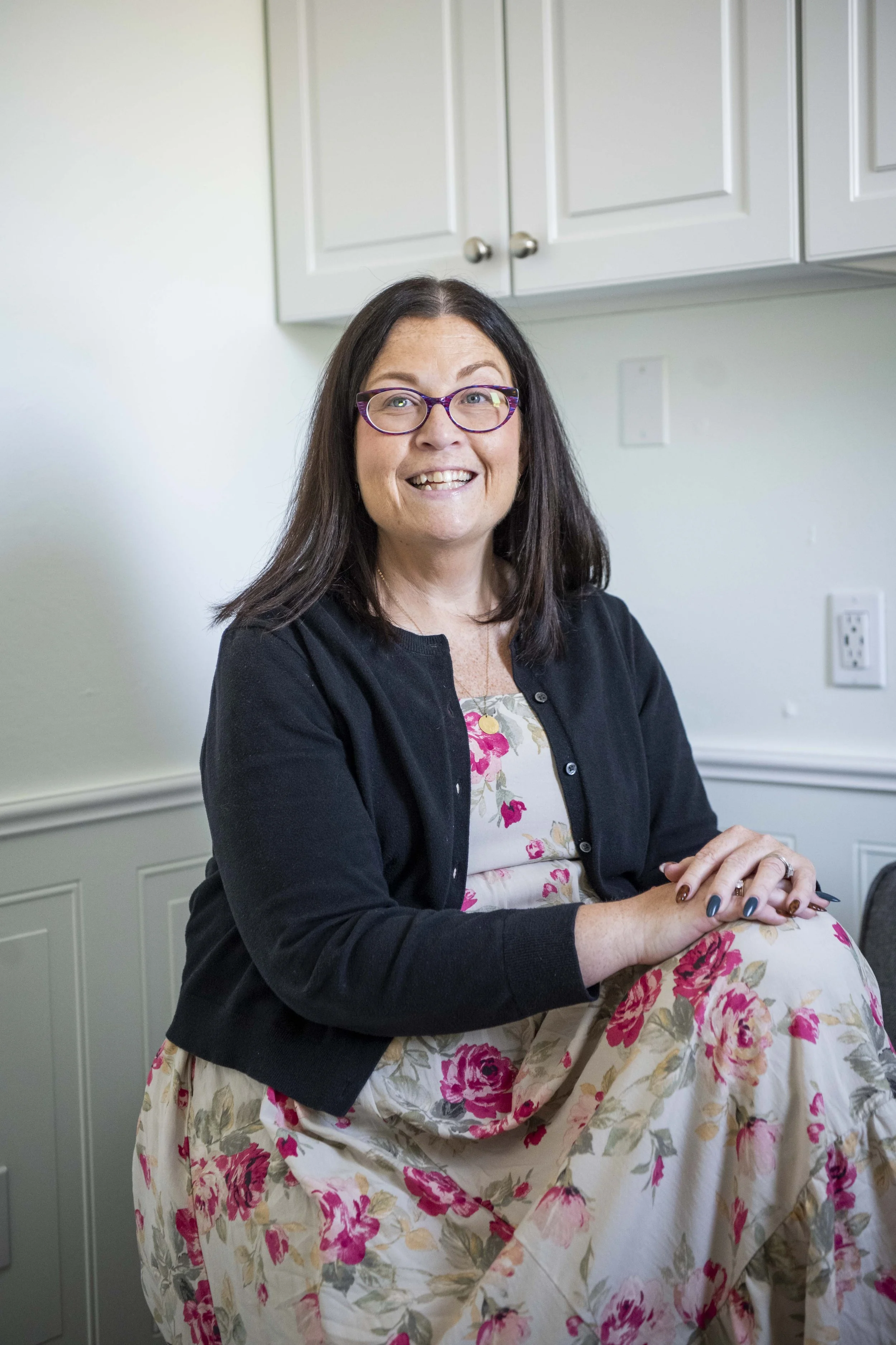 Lori McGrimmon sits in her office with one knee raised and hands resting on her knee while wearing a floral patterned dress