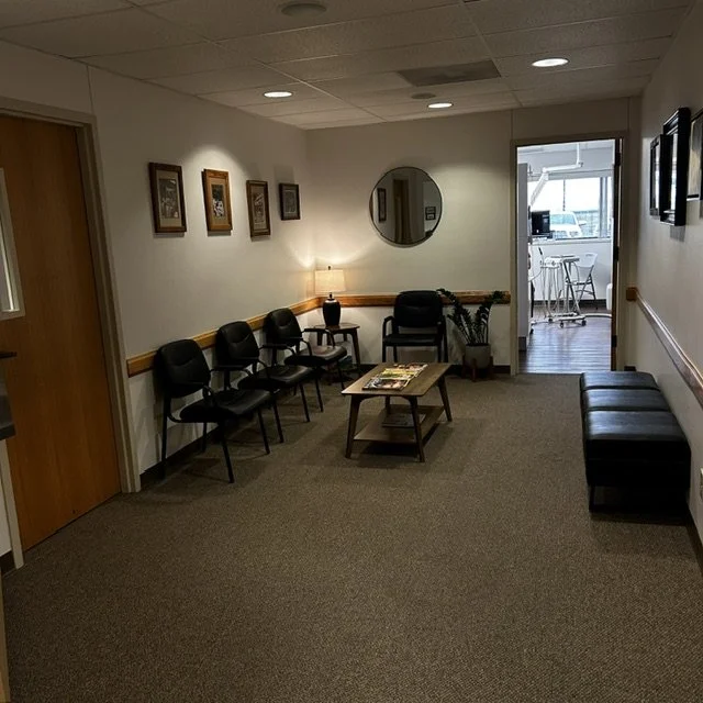 Empty waiting room with six black chairs against the wall, a small table with magazines, a black ottoman, a lamp, and a potted plant, leading to a kitchen area with a breakfast bar and stools.