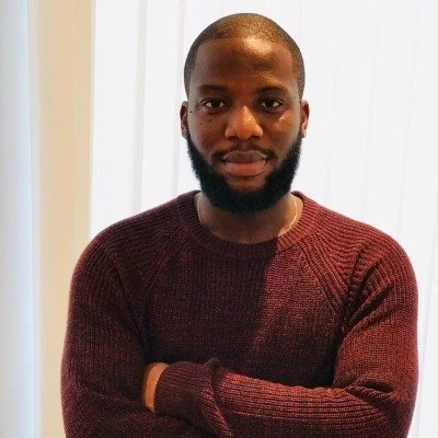 Man with a beard wearing a maroon sweater standing with arms crossed in front of a white curtain.