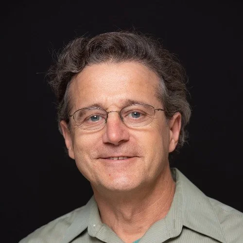 Smiling man wearing glasses and a green shirt against a dark background.