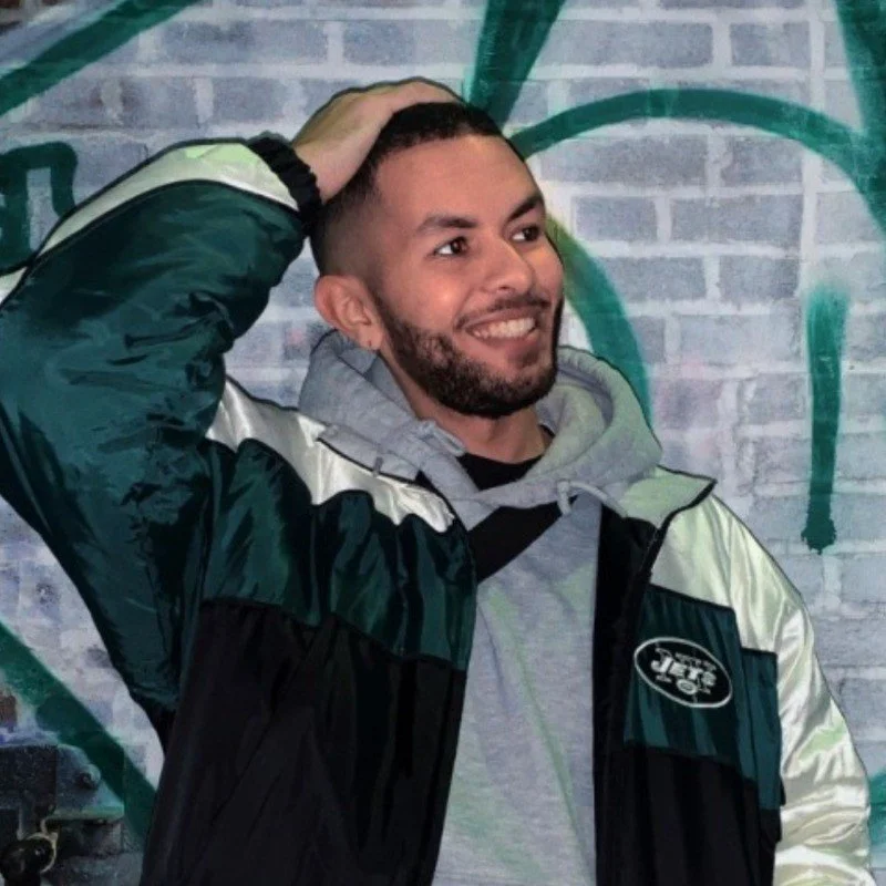 A young man with a beard smiling, wearing a Jets jacket and a gray hoodie, standing in front of a brick wall with green graffiti.