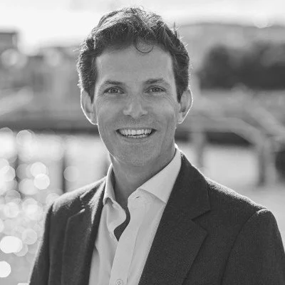 Black and white photo of a smiling man in a suit jacket and white shirt outdoors with blurred background