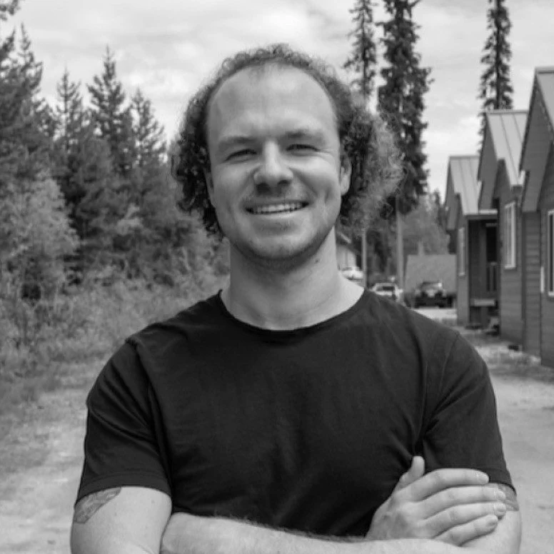 Smiling person with curly hair and crossed arms standing outdoors with trees and cabins in the background.