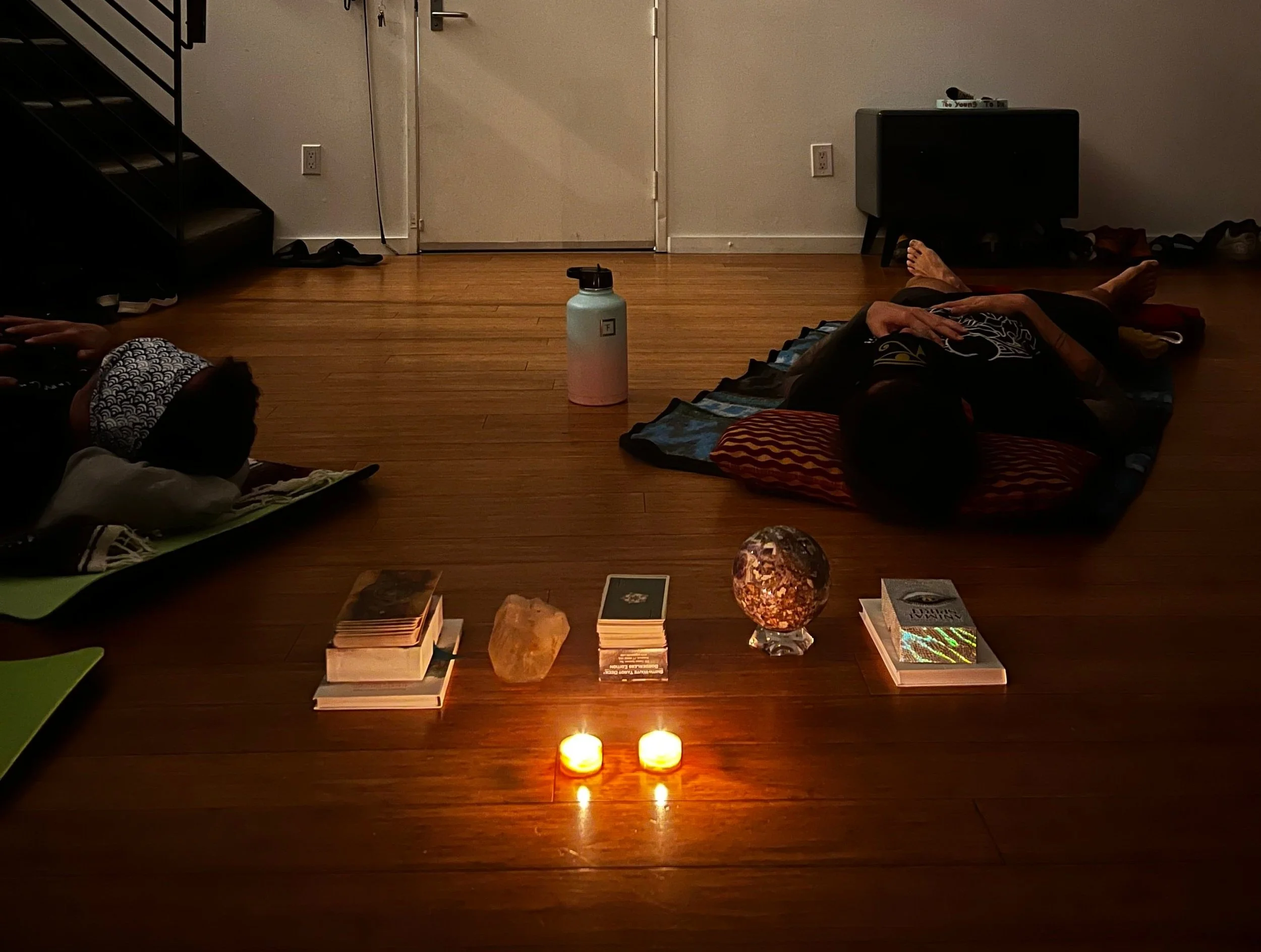 A dimly lit room with two individuals lying on mats, possibly meditating or relaxing. The room features a wooden floor and a central arrangement of candles, books, and a crystal sphere. A water bottle is visible in the background, near a door and a small cabinet.