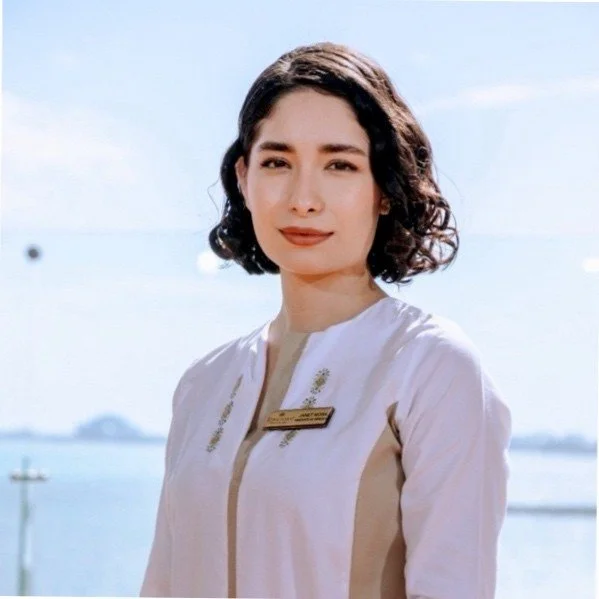 A young woman with short, curly dark hair and light skin, wearing a white uniform with a name tag, standing outdoors near water with a clear sky background.