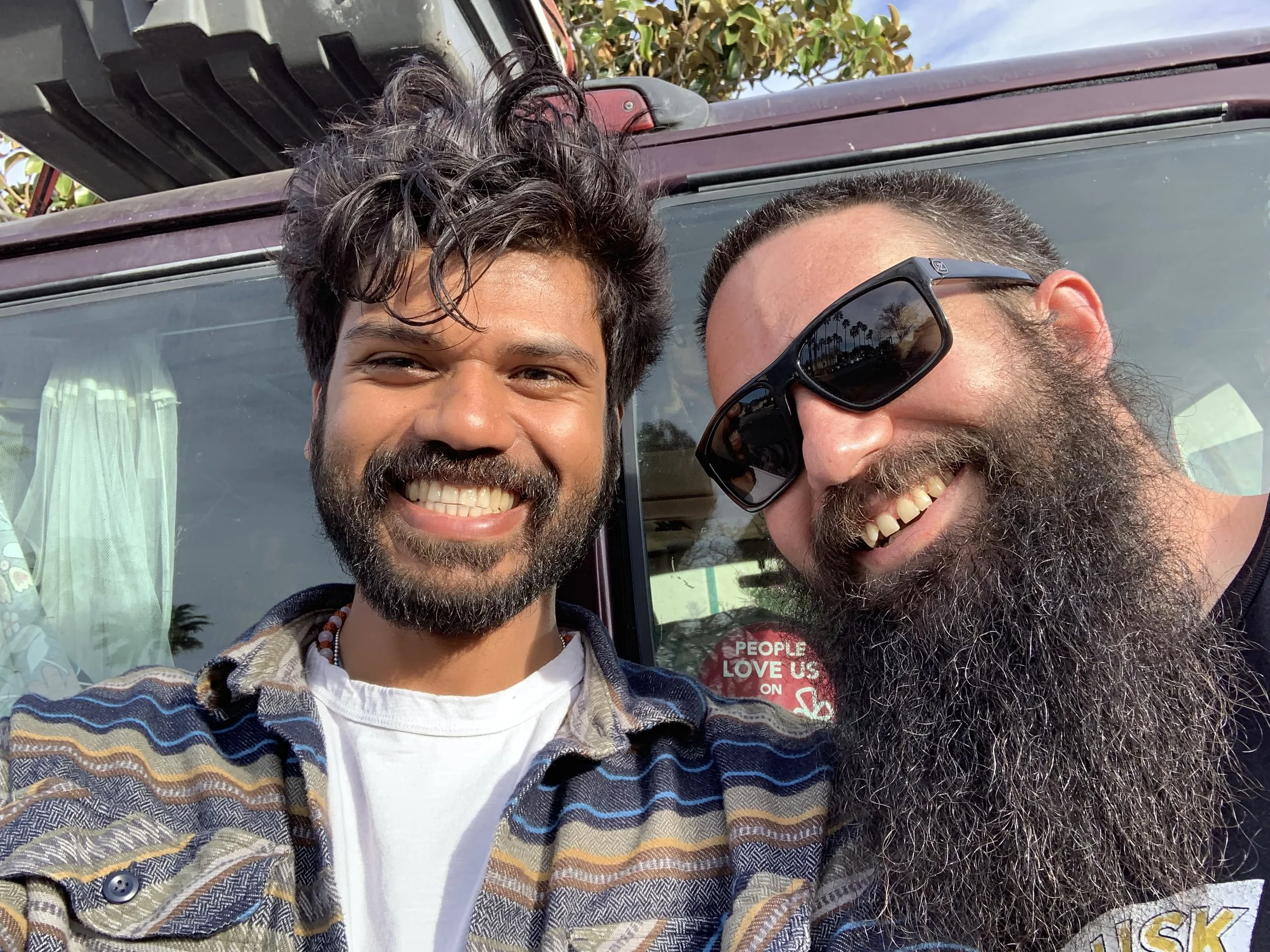 Two men smiling, one with sunglasses, standing outdoors next to a vehicle.