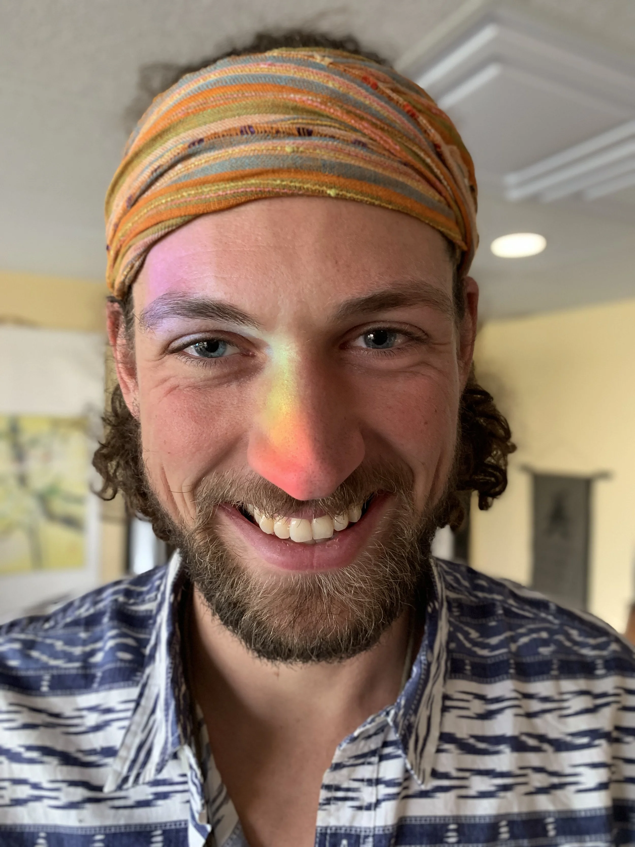 Smiling man wearing a patterned headband and shirt, with a rainbow reflection on his face.