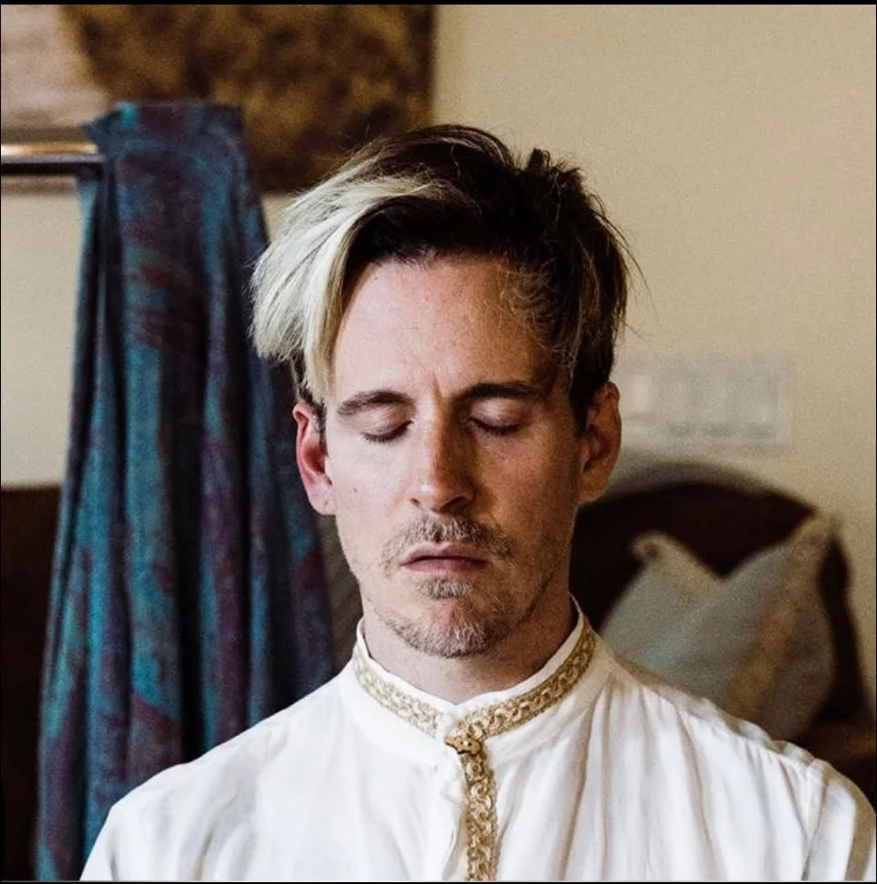 Person with closed eyes wearing a white shirt with gold embroidery.