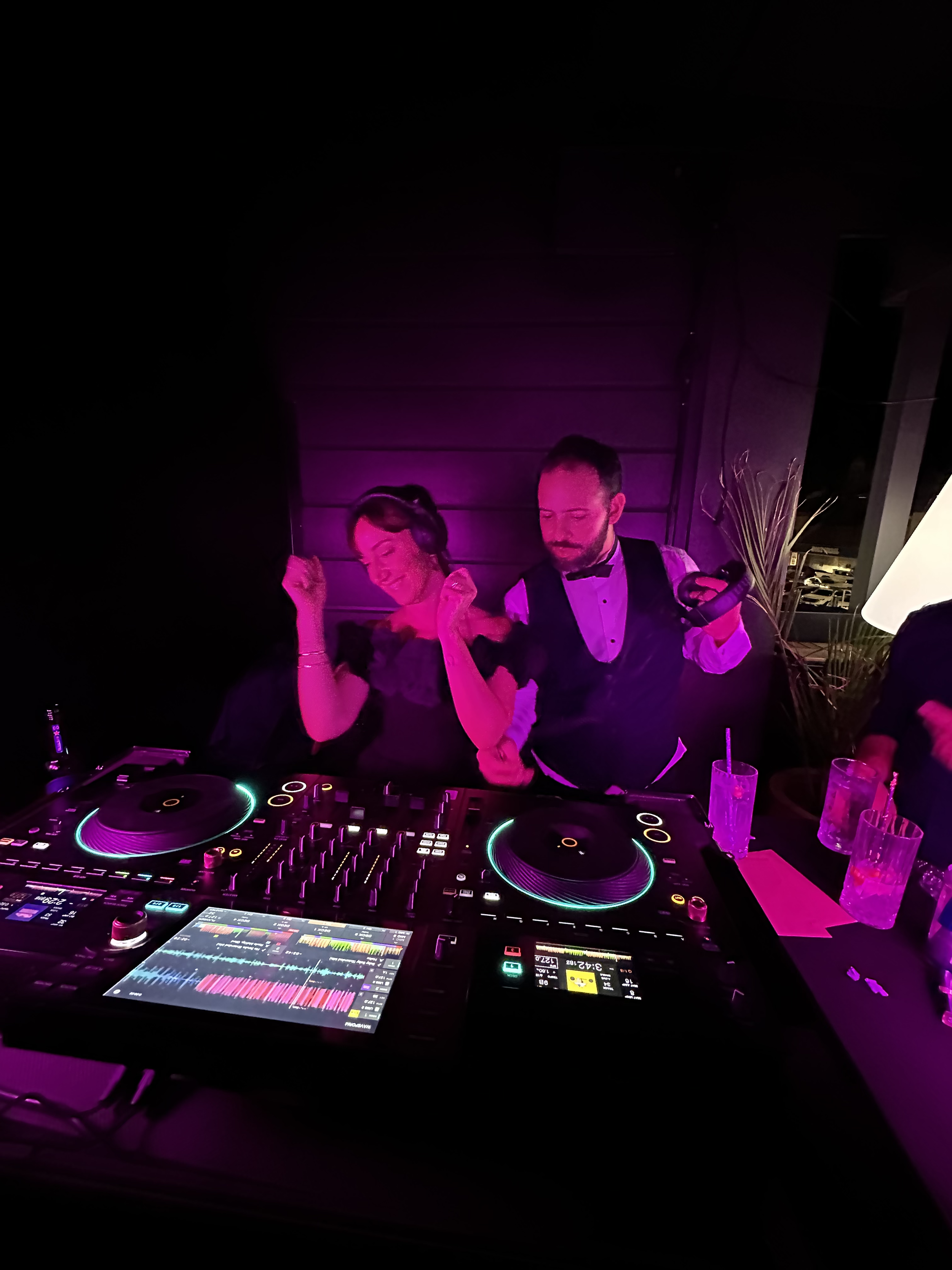 Man and woman dancing at a DJ booth with colorful lights and drinks on the table at an indoor party.