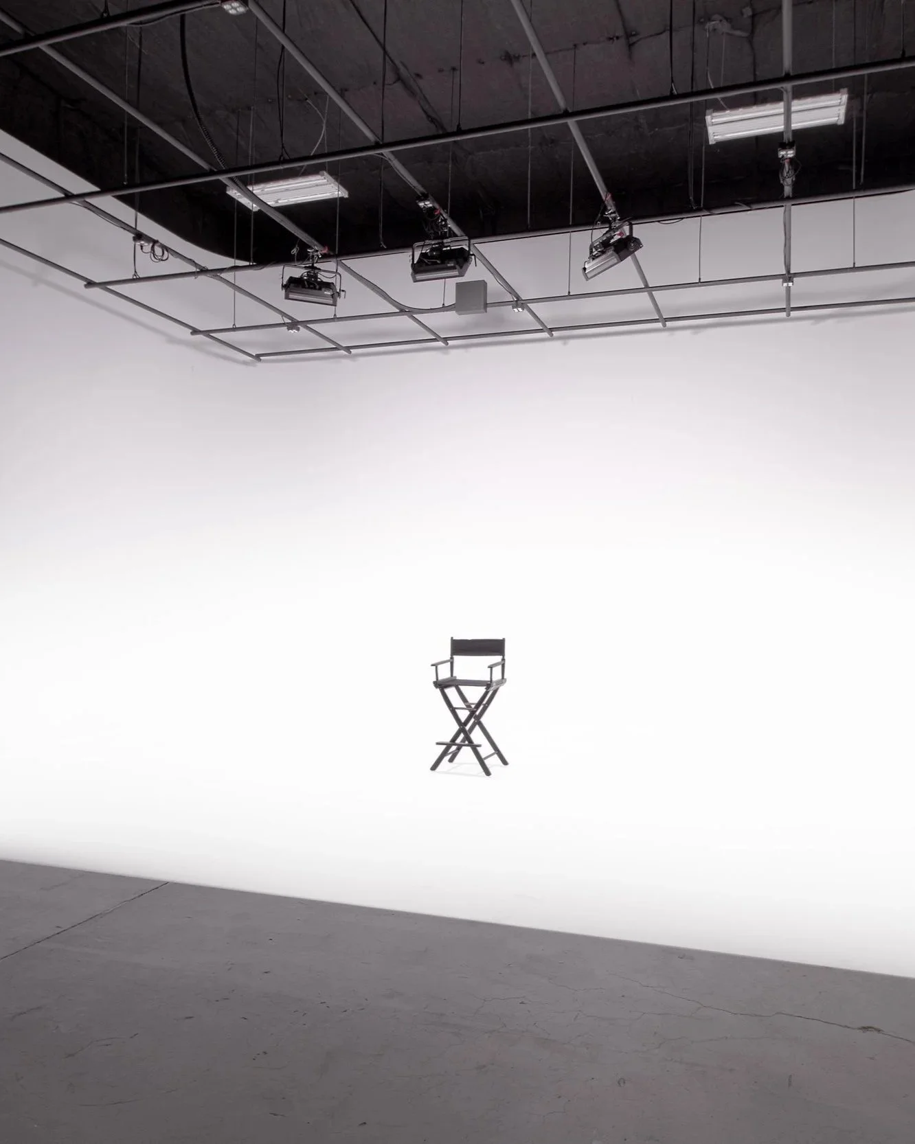 An empty director's chair hangs in a photography studio with a white backdrop, overhead lights, and studio equipment.