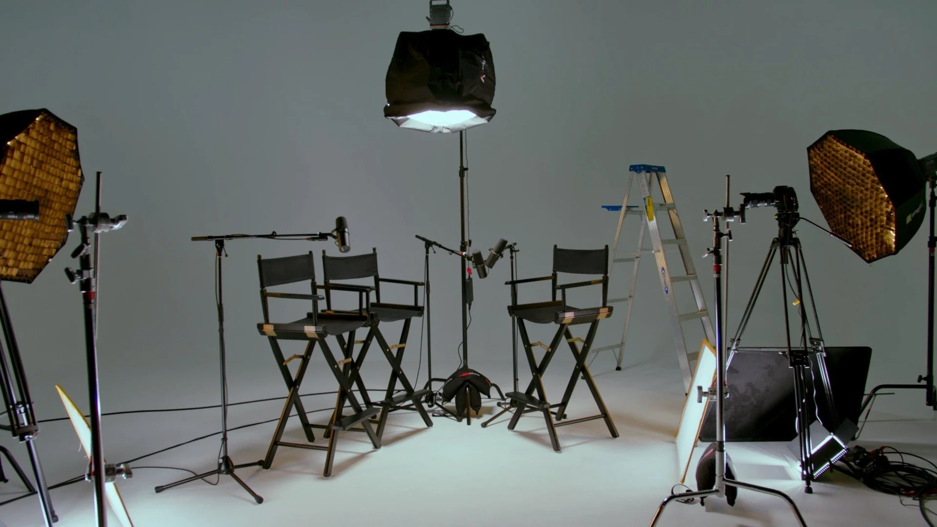 Film or interview recording set with two director chairs, studio lights, microphones, a ladder, and a camera on a tripod in a gray studio background.