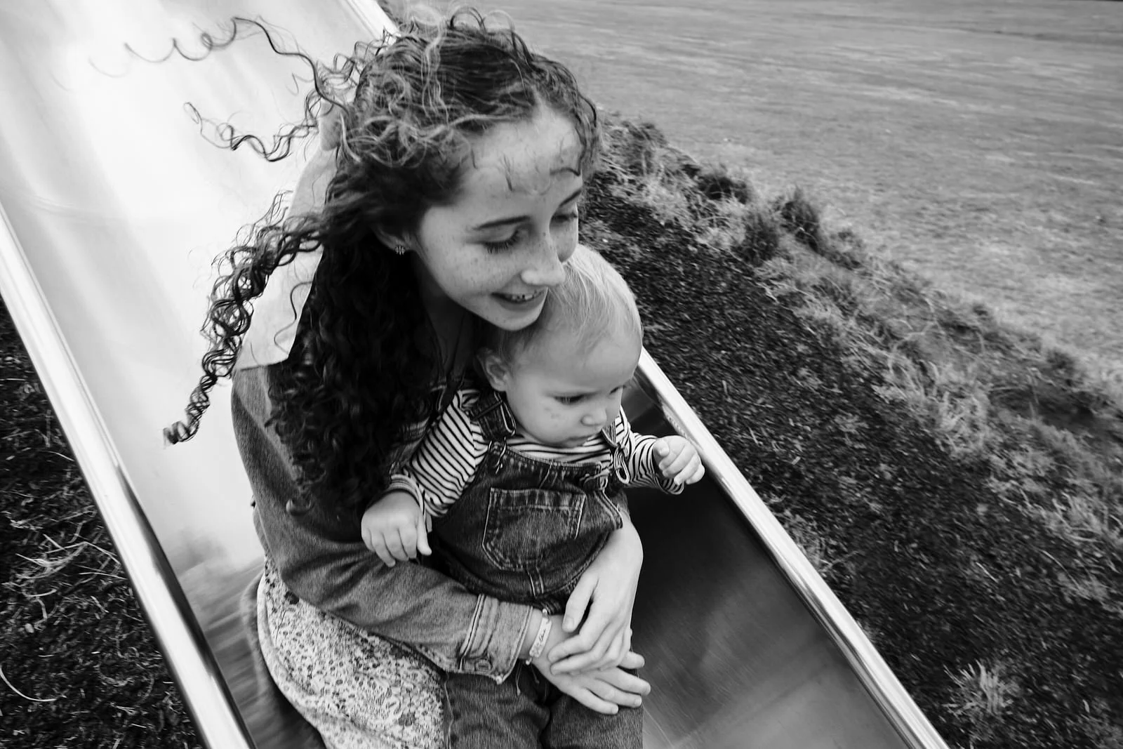 A girl with curly hair and a boy with short hair sliding down a playground slide outdoors on a cloudy day.