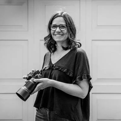 Woman with glasses smiling, holding a camera, standing in front of a garage door.