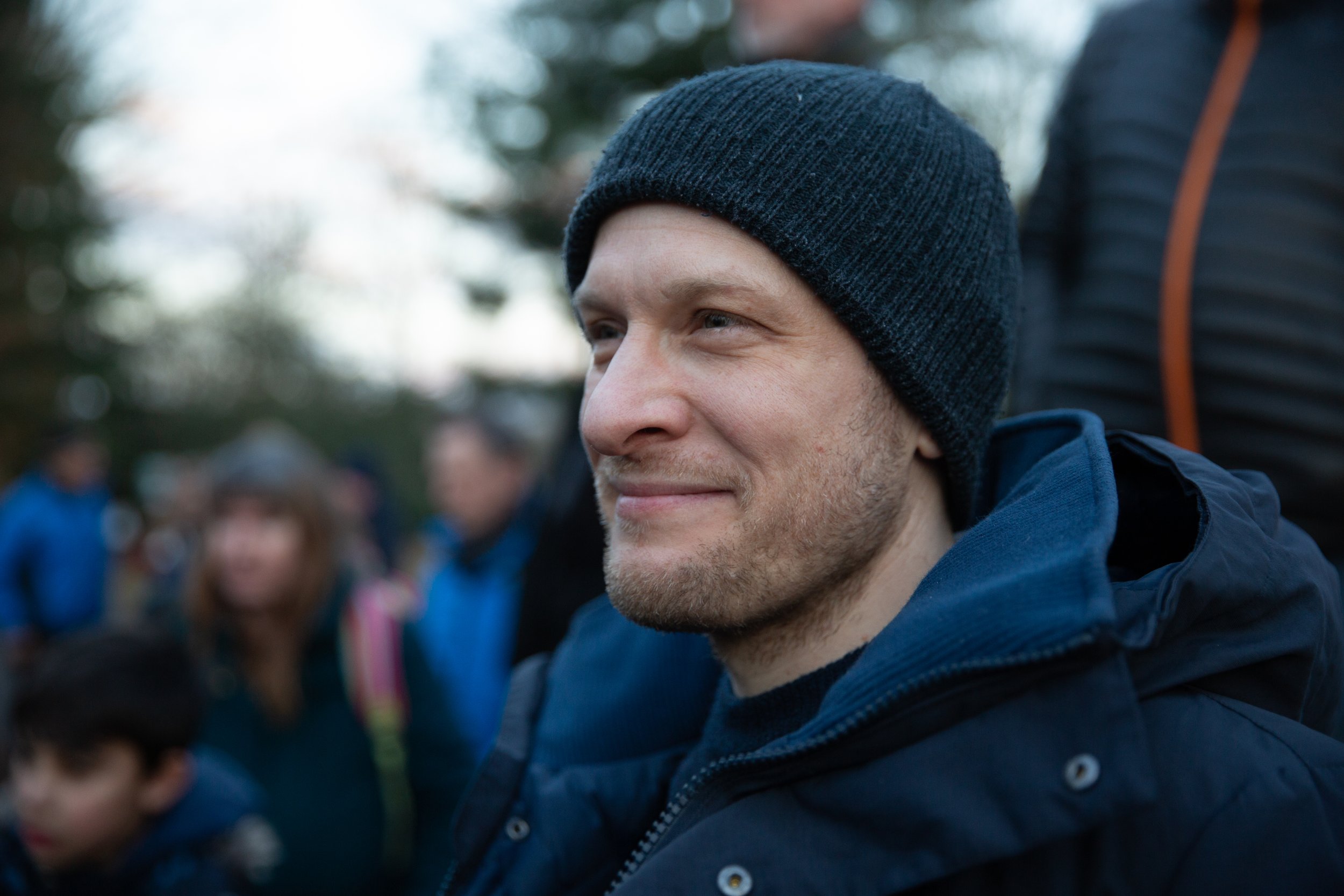 Close-up of a smiling man wearing a dark beanie and jacket, outdoors, with a blurred crowd in the background.