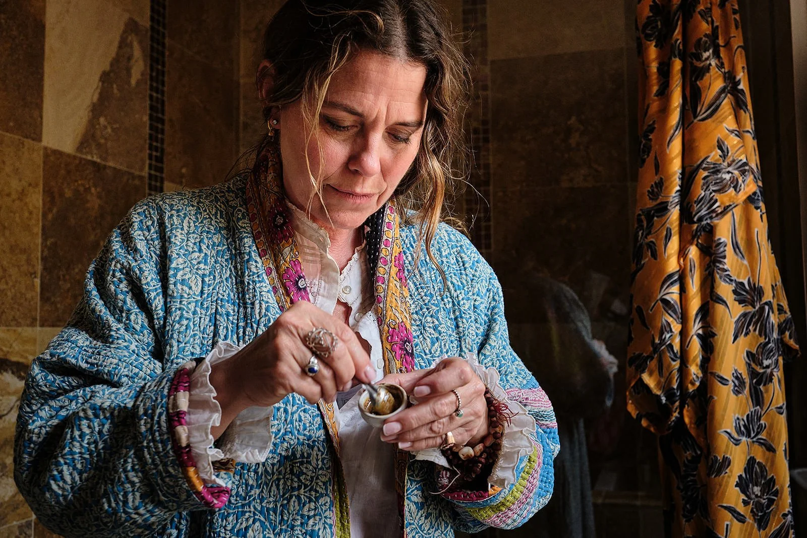 A woman with wavy hair wearing a multicolored, patterned jacket and jewelry, looking down while scooping ice cream from a small container.