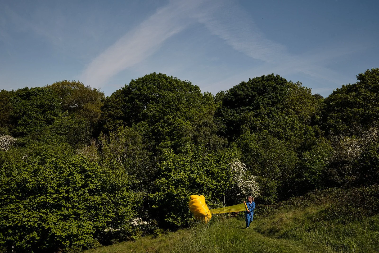 A person wearing blue overalls flying a yellow kite in a green outdoor area with trees and a blue sky.