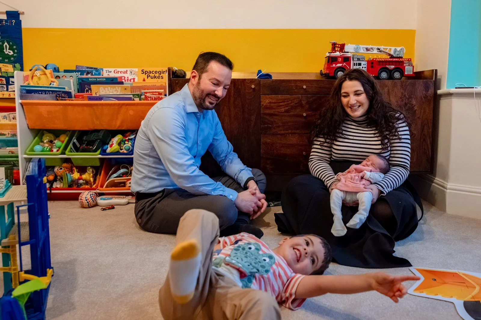 A family sitting on a carpeted floor in a playroom. A woman holds a baby, a young boy points and lies on his back, and a man sits nearby. The room has shelves filled with toys and books, including a fire truck on the cabinet.