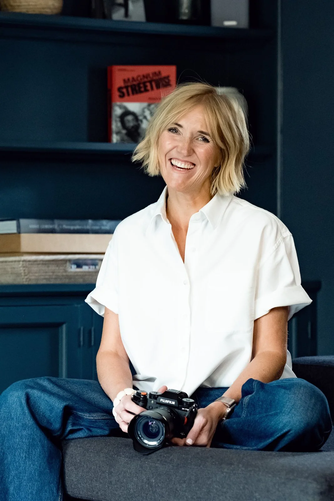 A woman with blonde hair, wearing a white shirt and blue jeans, smiling and holding a camera, sitting on a dark sofa in a modern living room.