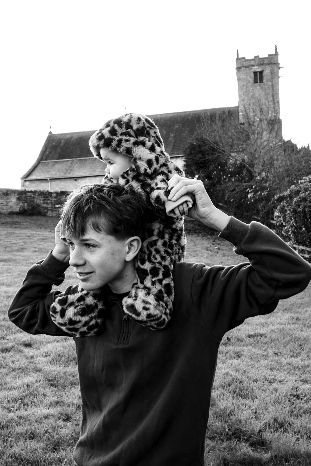 A young man carrying a small child on his shoulders outdoors in front of a church or old building, both dressed in warm clothing, with trees and grass in the background, black and white photo.