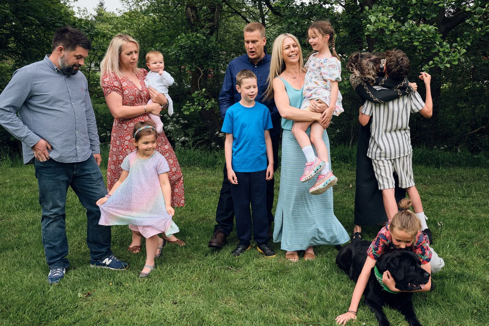Family gathering outdoors with multiple adults and children on a grassy area surrounded by trees, some children playing and hugging, and a black dog on a leash.