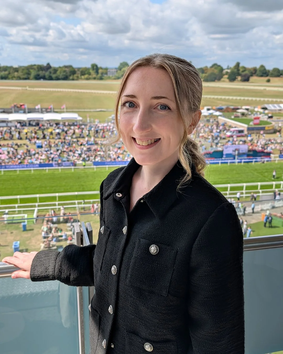 A woman with light brown hair smiling at the camera, standing at a racetrack with a crowd of people and the racecourse in the background.
