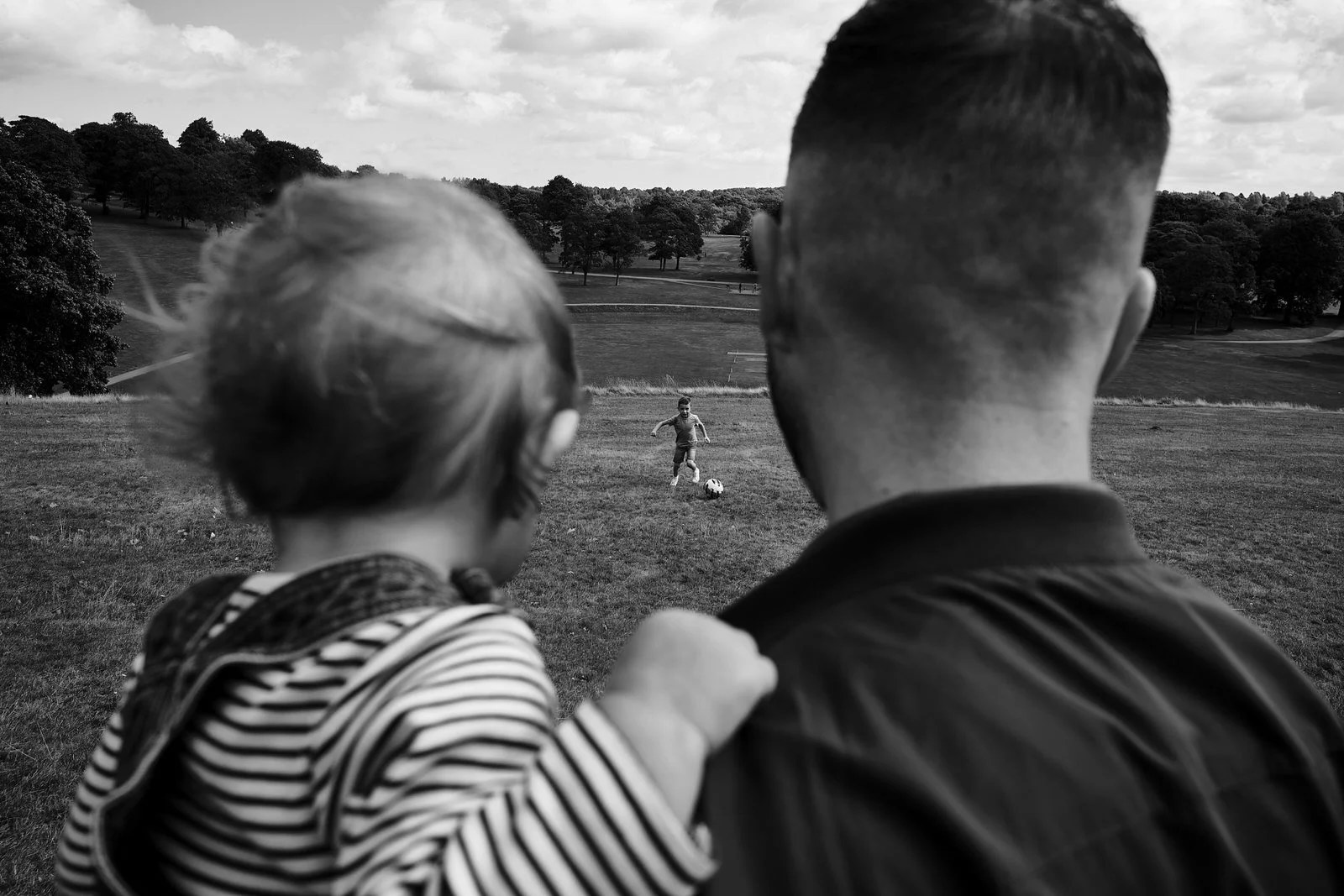 A man and a young girl watching a boy playing soccer on a field in a park with trees and cloudy sky in the background.